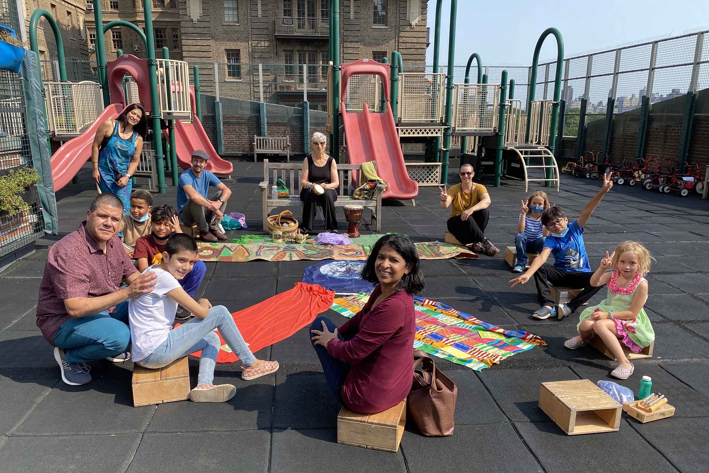 A diverse group of children and adults sit on a colorful cloth on a rooftop playground, posing for the photo with smiles and peace signs, with slides and green playground equipment behind them.